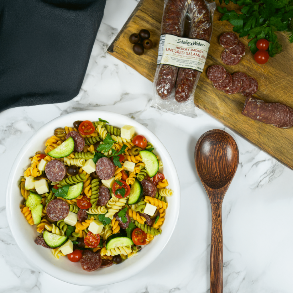 Overhead view of a colorful pasta salad with salami, zucchini, tomatoes, olives, and cheese cubes next to packaging on a marble surface
