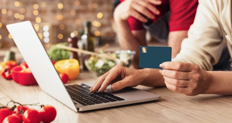 Hands holding a credit card while shopping for food online on a laptop in a kitchen